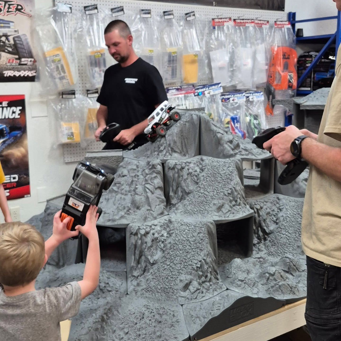 People operating remote control trucks on a rocky, gray terrain display in a hobby store.