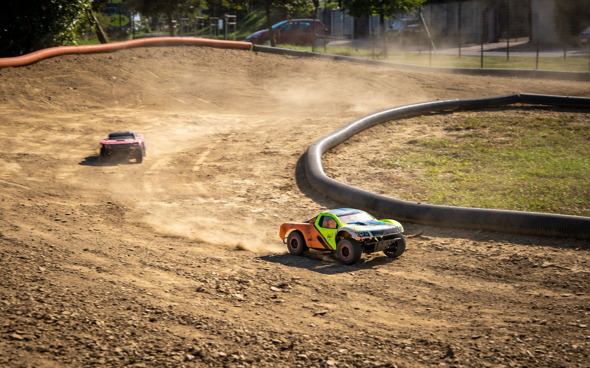 Two remote-controlled trucks racing on a dirt track, kicking up dust; green and orange truck in foreground.