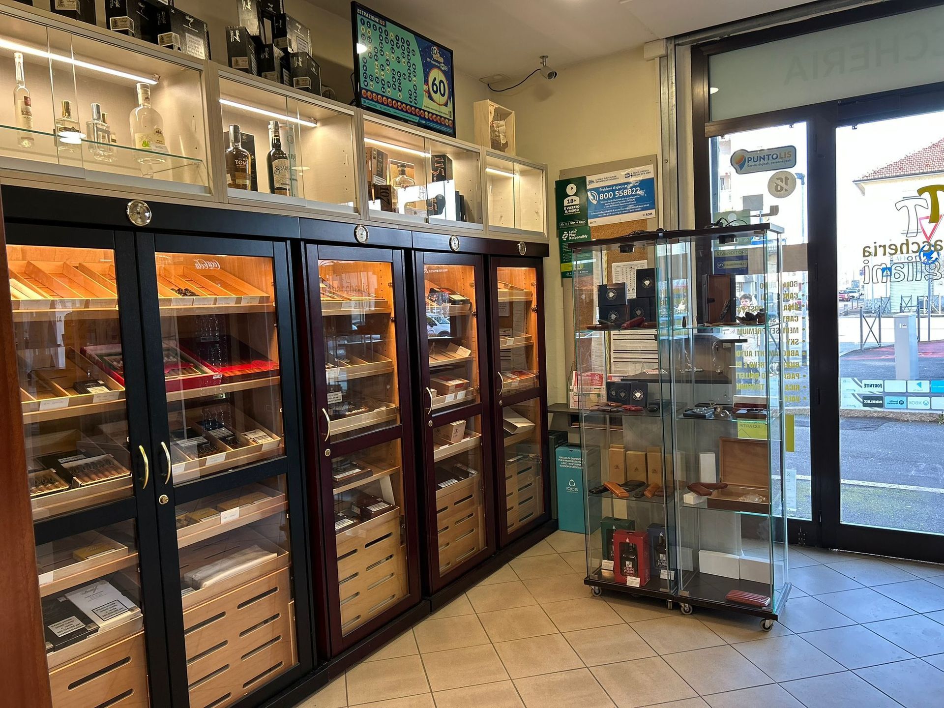 Interno di un negozio di sigari con humidor che espongono i sigari, bottiglie di liquore sugli scaffali e una vetrina vicino a una finestra.