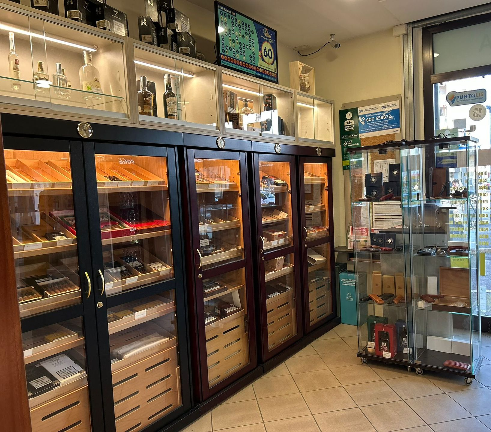 Interno di un negozio di sigari con humidor, scaffali illuminati che espongono sigari e liquori. Vetrine vicino alla finestra.