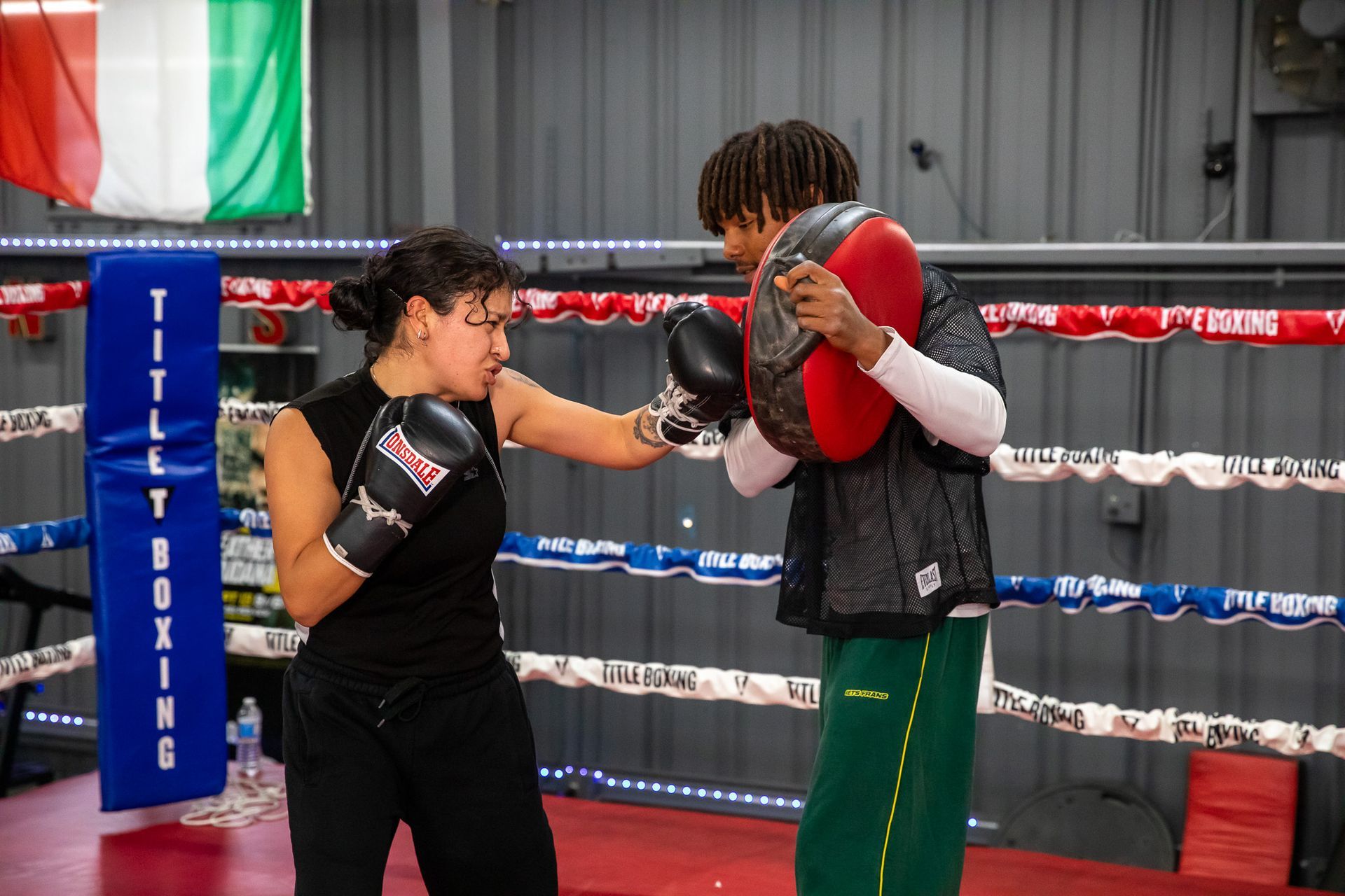 Woman boxing with trainer in a ring; Italian flag, black and green workout attire, punching focus.