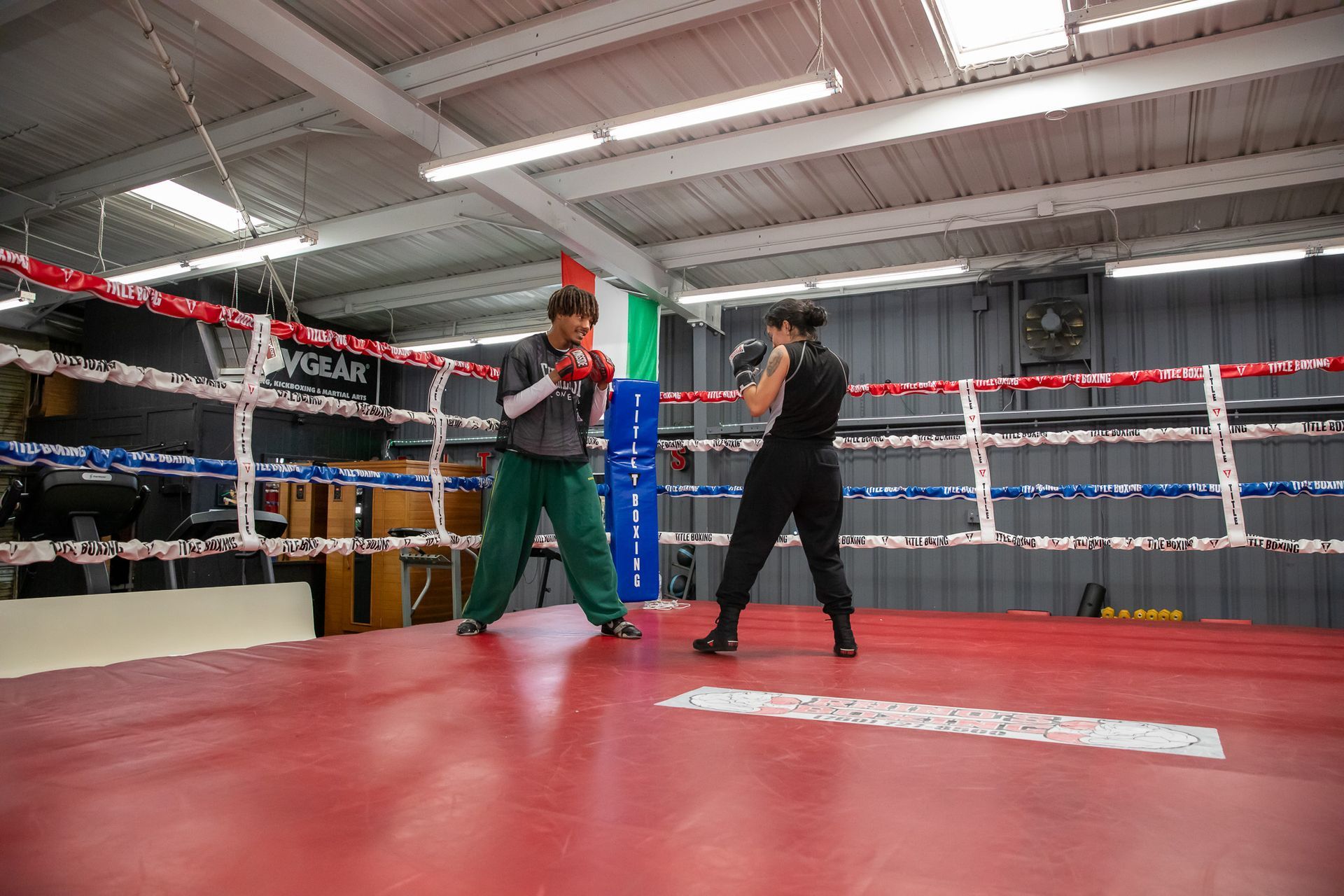 Two women boxing in a red ring; one in green pants, one in black, practicing punches.
