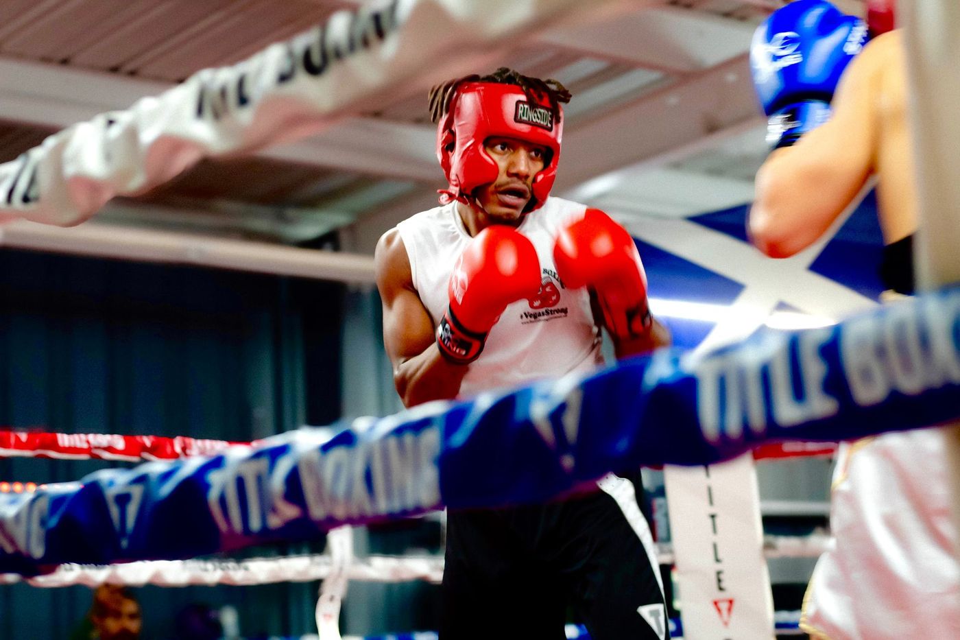 Boxer in red headgear and gloves faces opponent in blue gloves, inside a boxing ring.