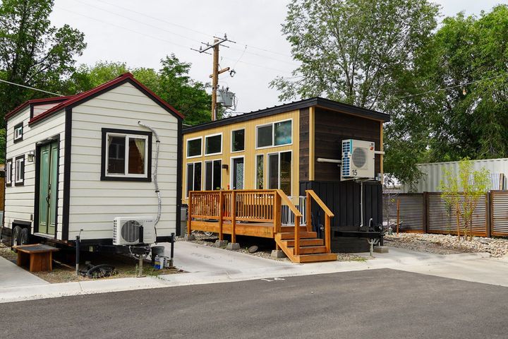 Two tiny houses on trailers parked on a concrete surface. One is white with a red roof, the other is brown with a deck.