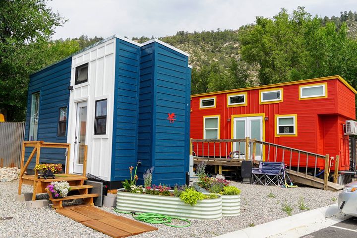 Two colorful tiny homes: a blue and white one and a red one, set in a gravel yard with mountains in the background.