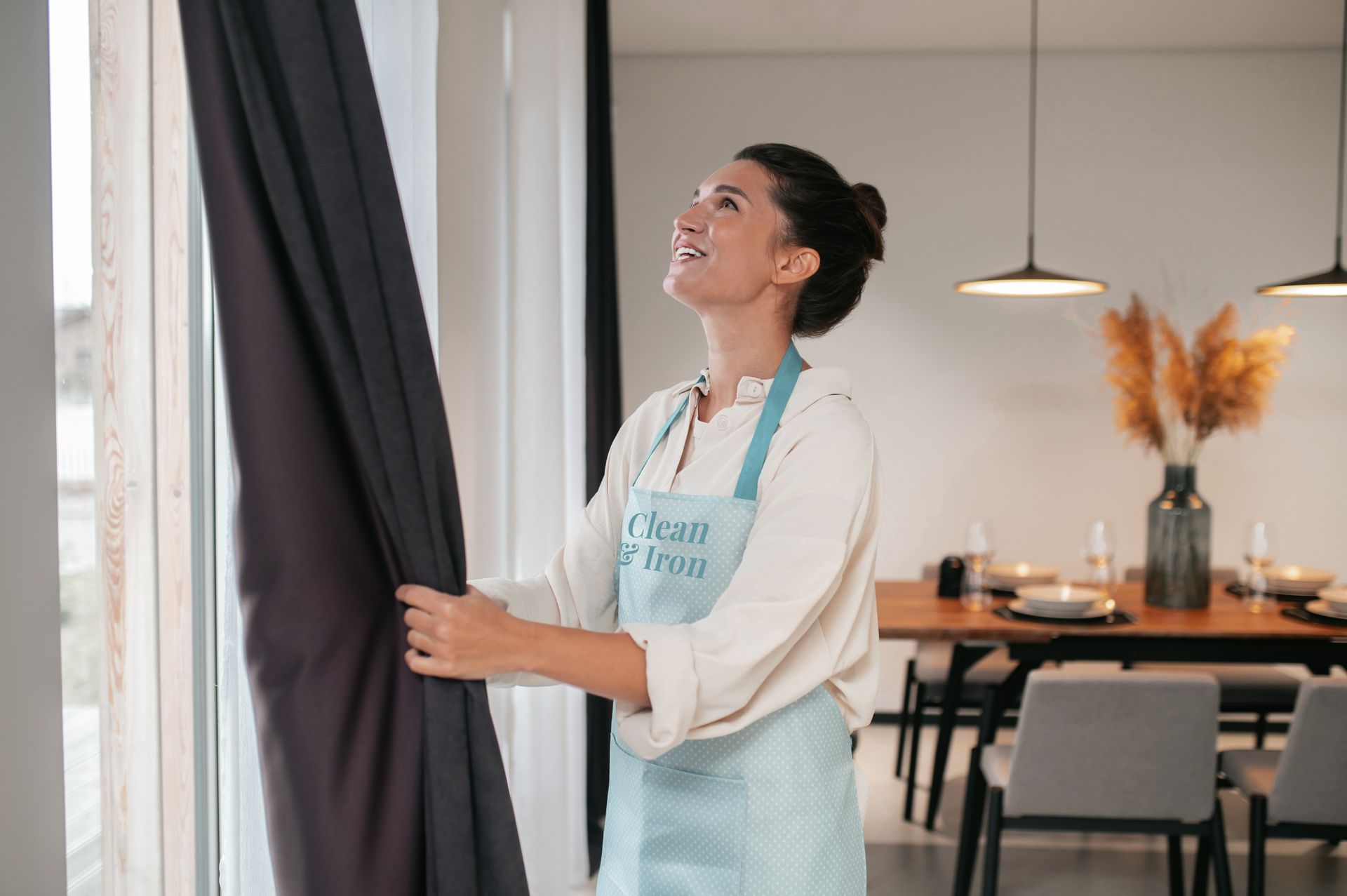 Dona amb davantal de clean and iron retirant cortines, somrient, en un menjador modern.