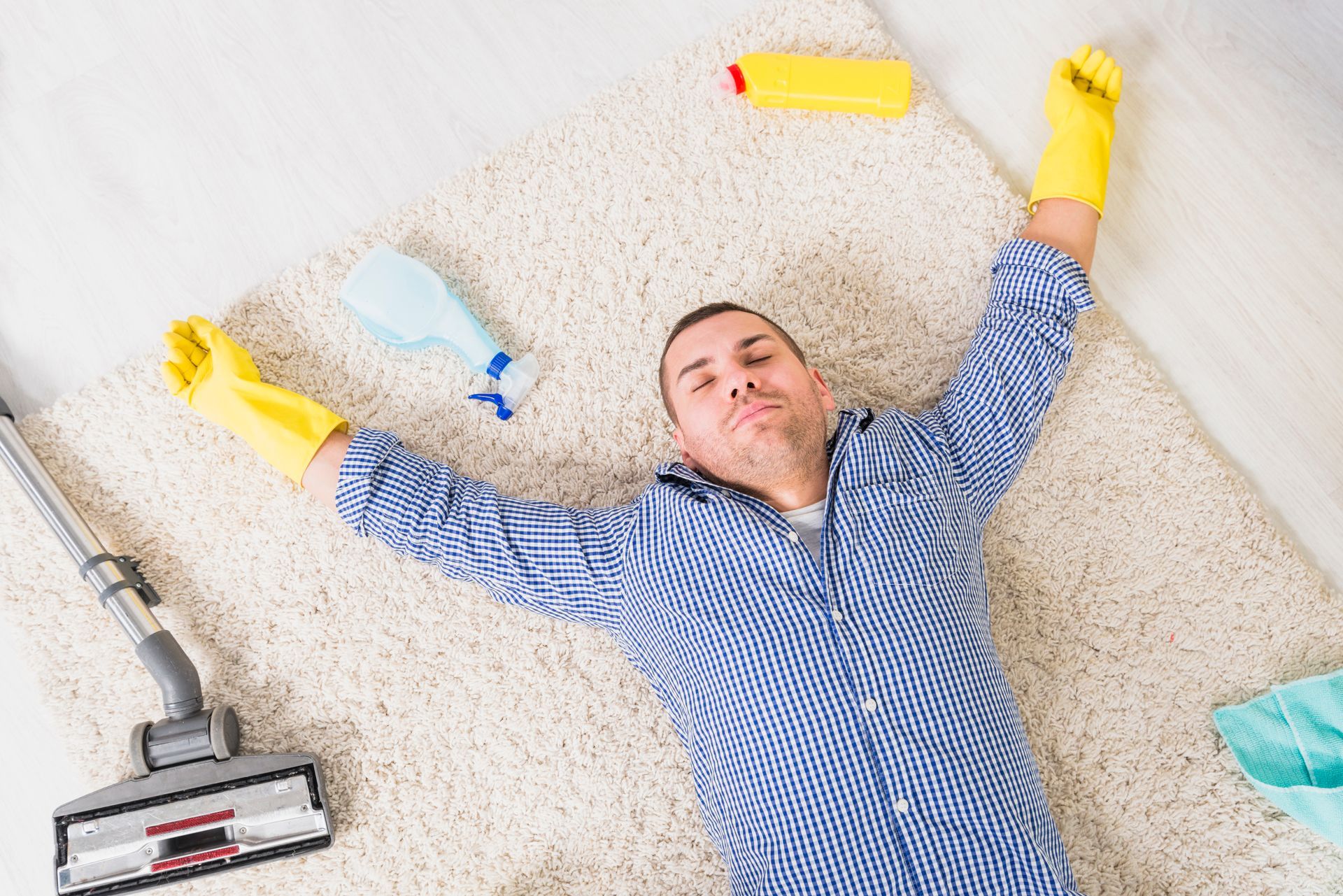 Hombre acostado exhausto sobre una alfombra después de limpiarla; usando guantes amarillos, rodeado de productos de limpieza.