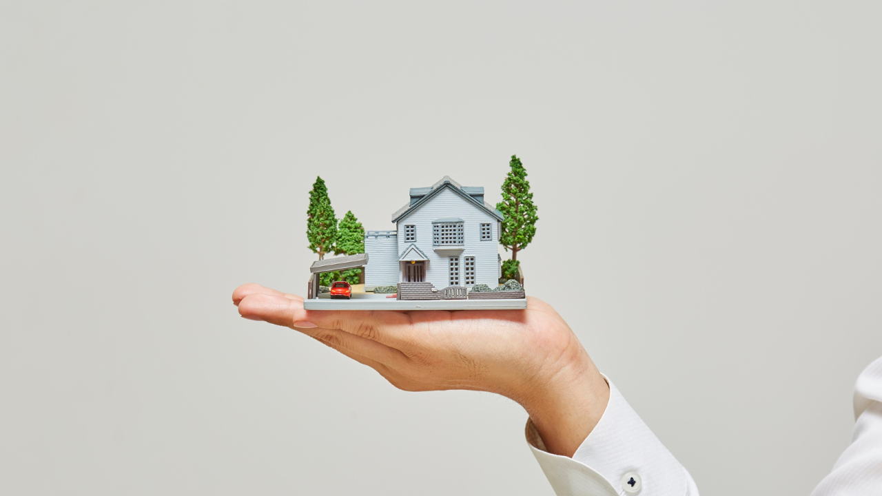 Hand holding a miniature house with trees; light background.