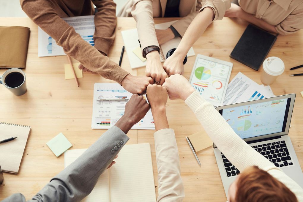 People fist bump over a desk with documents, a laptop, and a coffee cup.