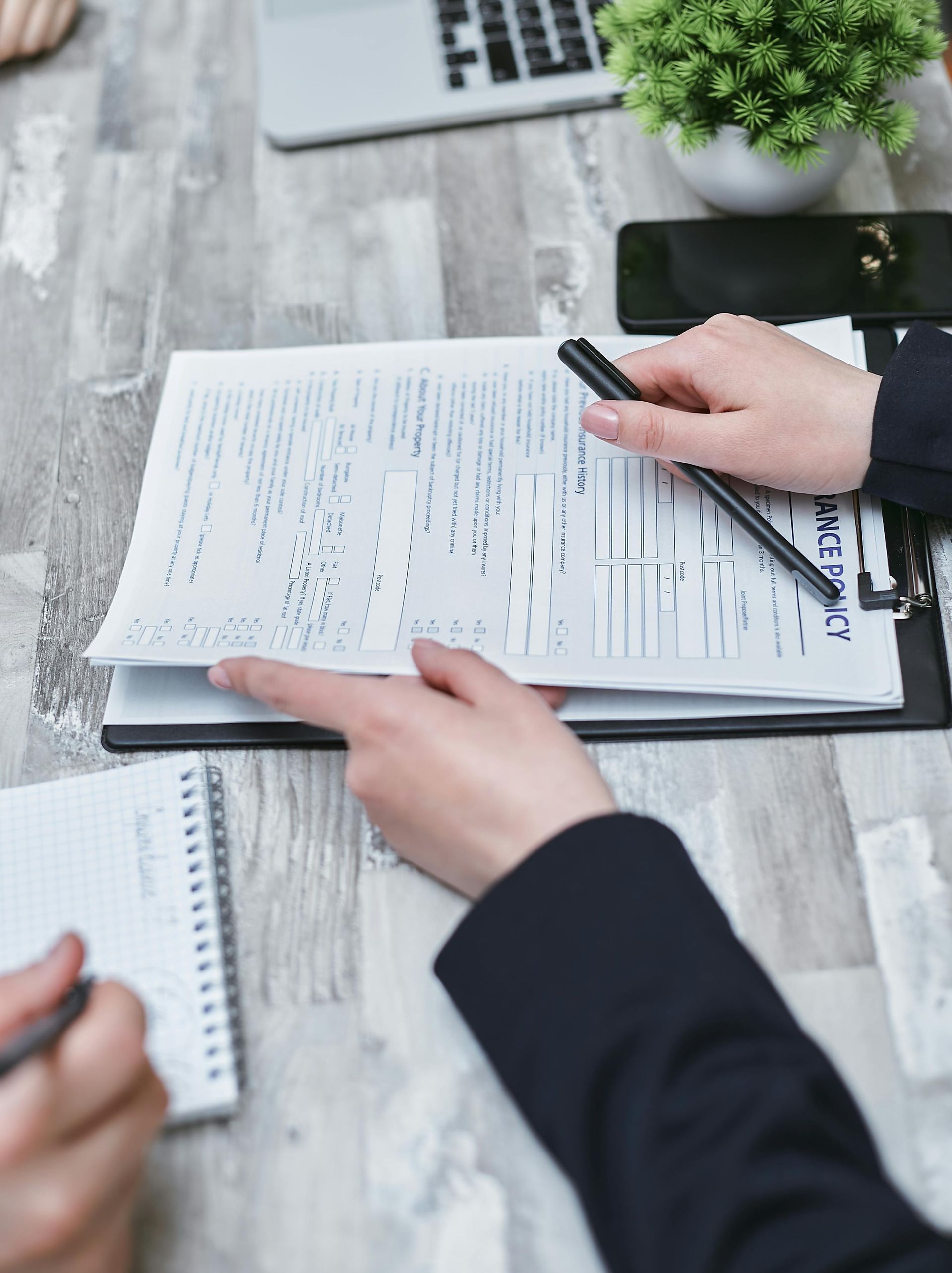 Hands holding a pen and pointing at a document on a desk, with a notepad and laptop nearby.