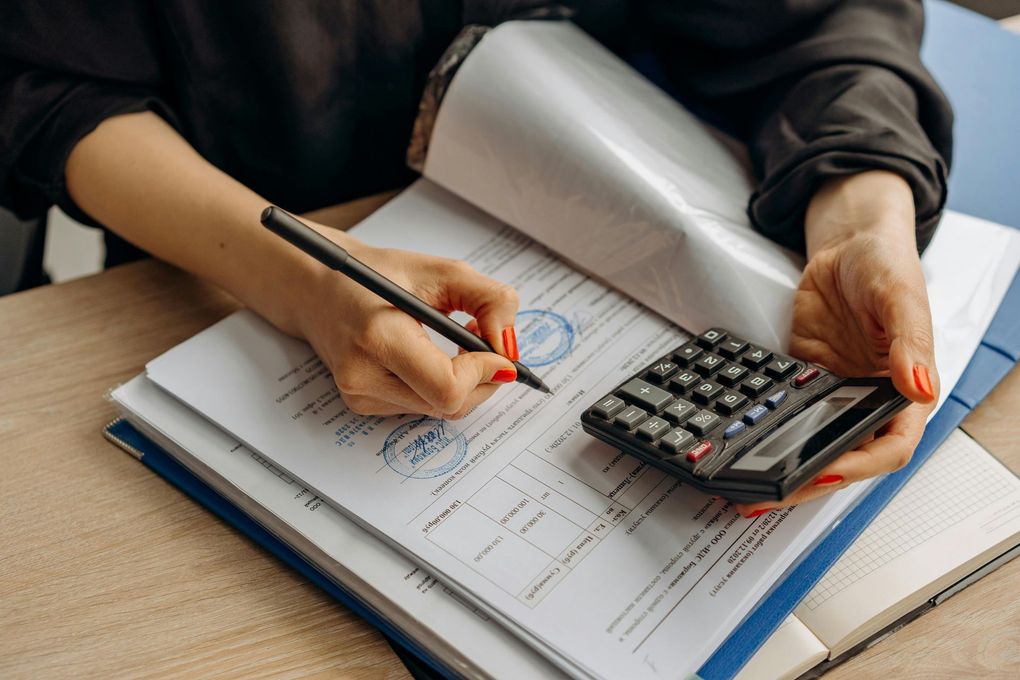 Person using a calculator while writing on paperwork. A seal stamp and notebook are nearby.