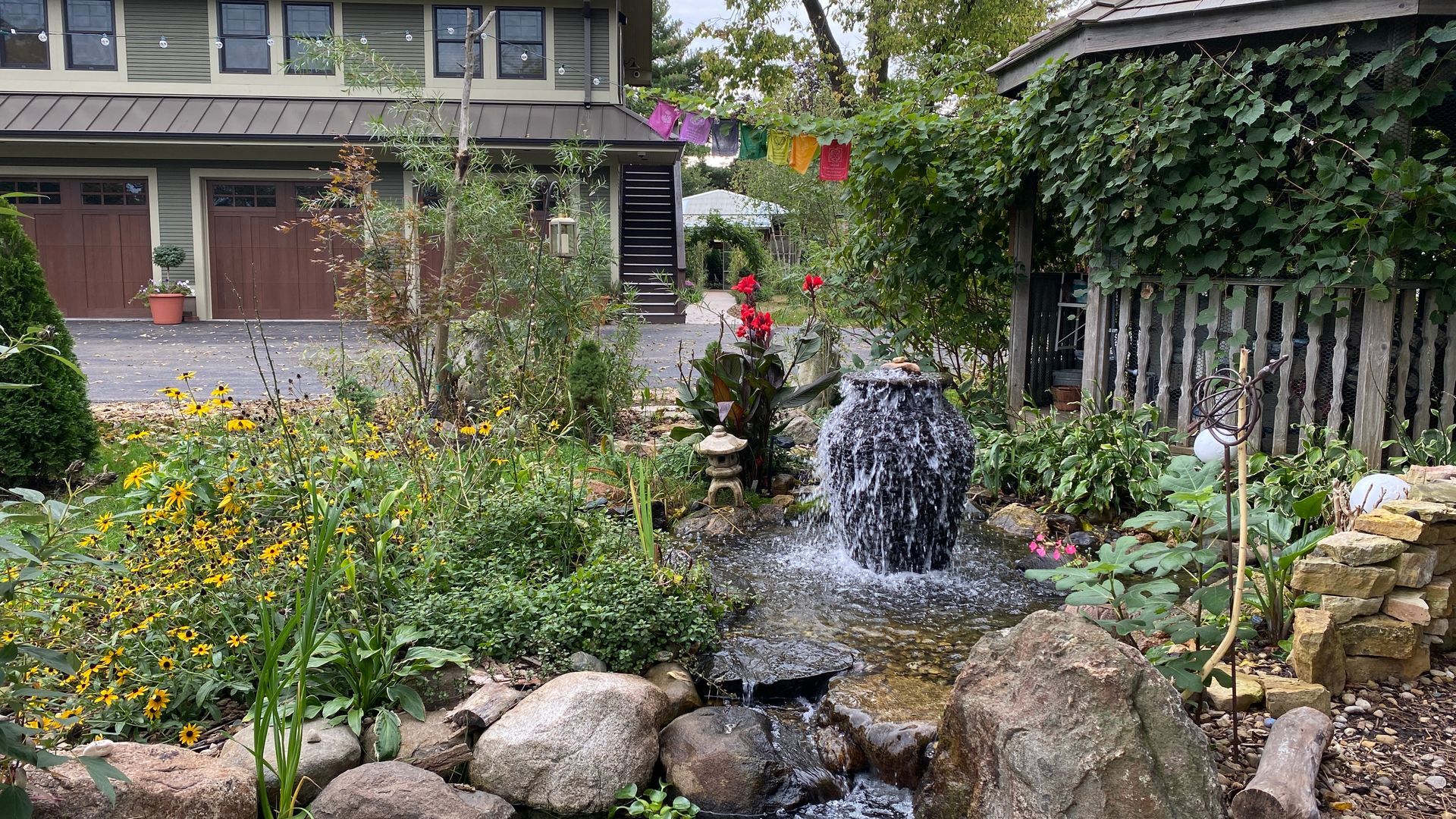 A waterfall is in the middle of a garden in front of a house.