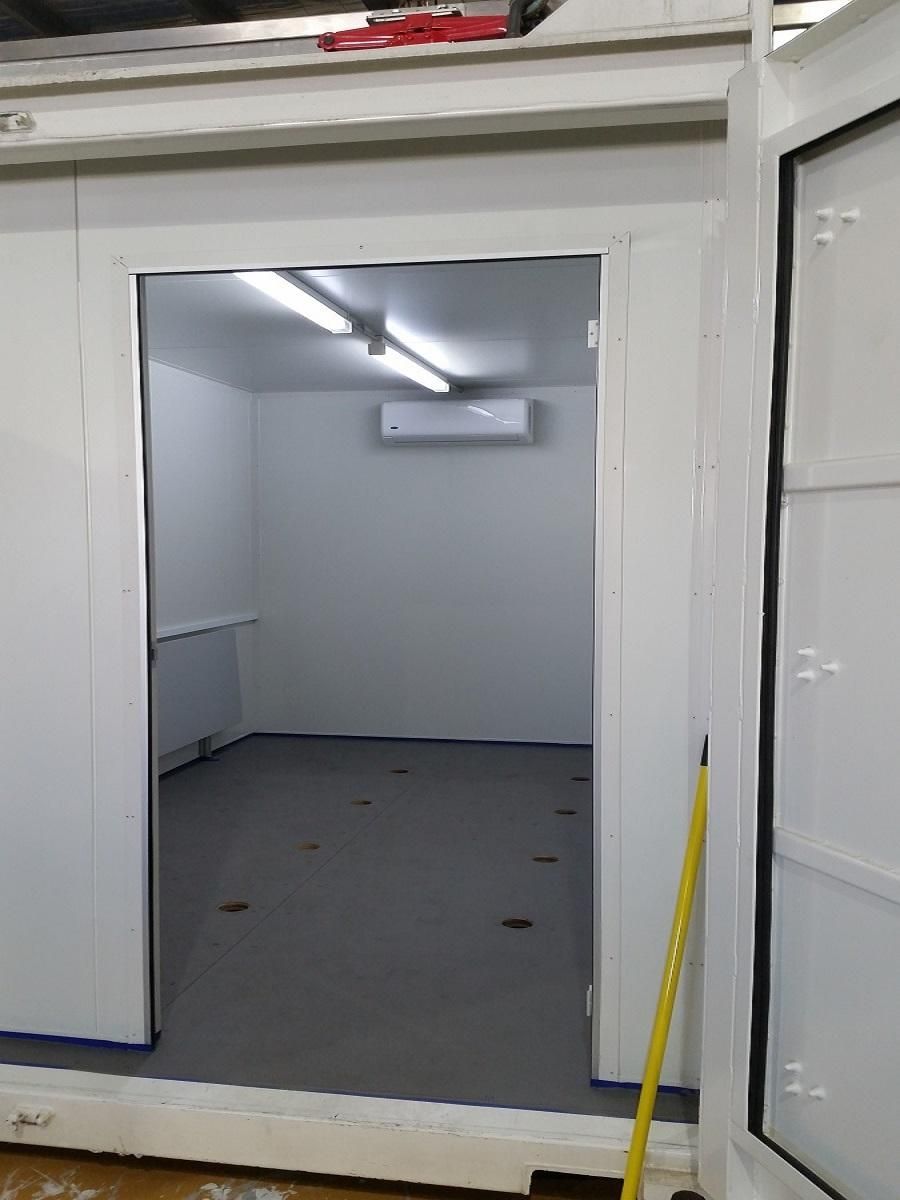 Interior of a white room with open door. Gray floor, AC unit on wall, fluorescent lights.