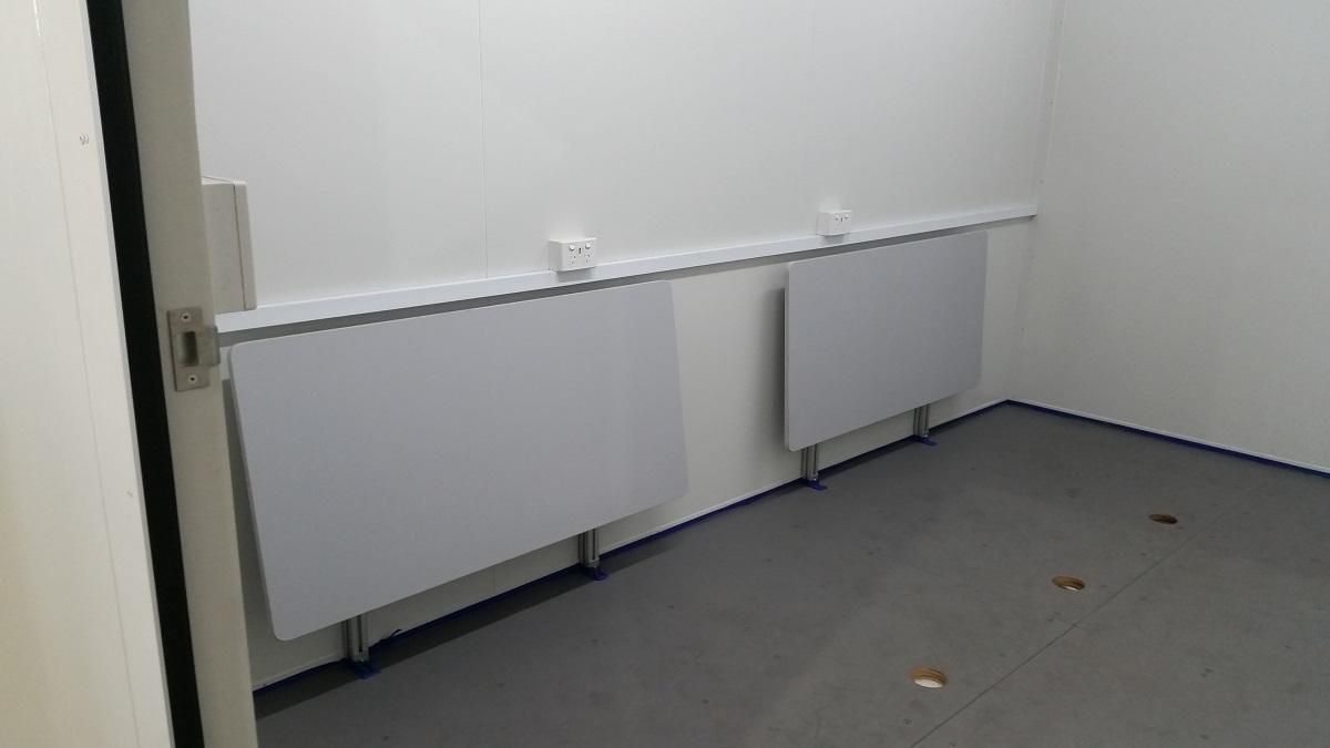 Two white folding tables mounted on a white wall in a room with gray carpet.