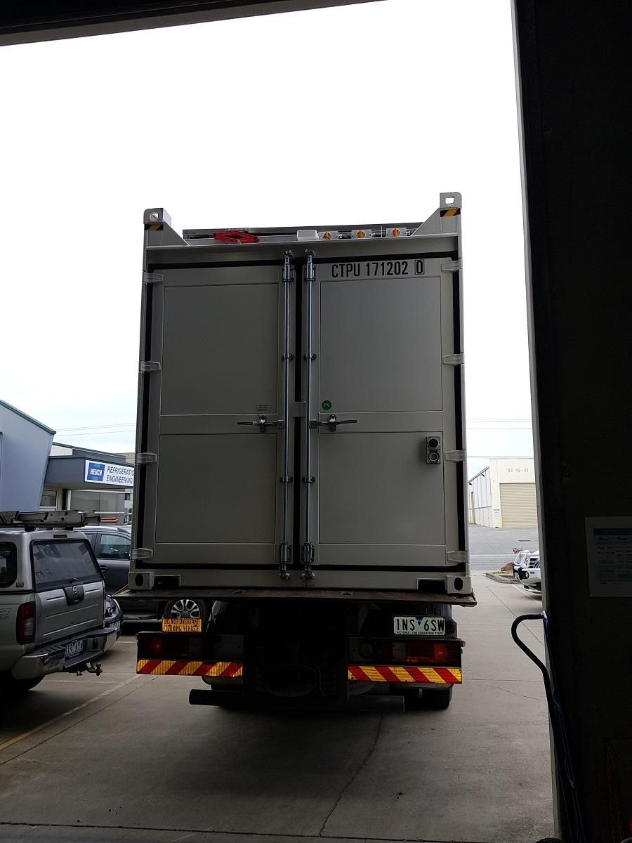 Rear view of a truck carrying a closed white shipping container, parked in a covered area.