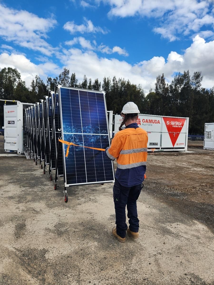 Worker in high-vis vest secures a stack of solar panels on a construction site with storage containers in the background.