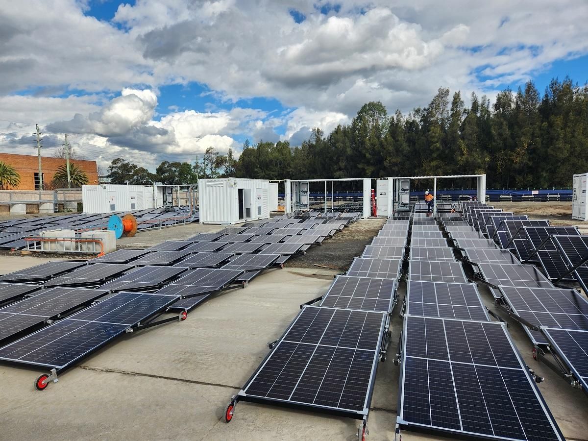 Solar panels laid out on concrete, with shipping containers in the background, under a cloudy sky.