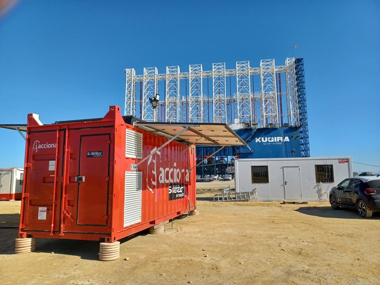 Red container with logo next to large blue structure under a clear sky.