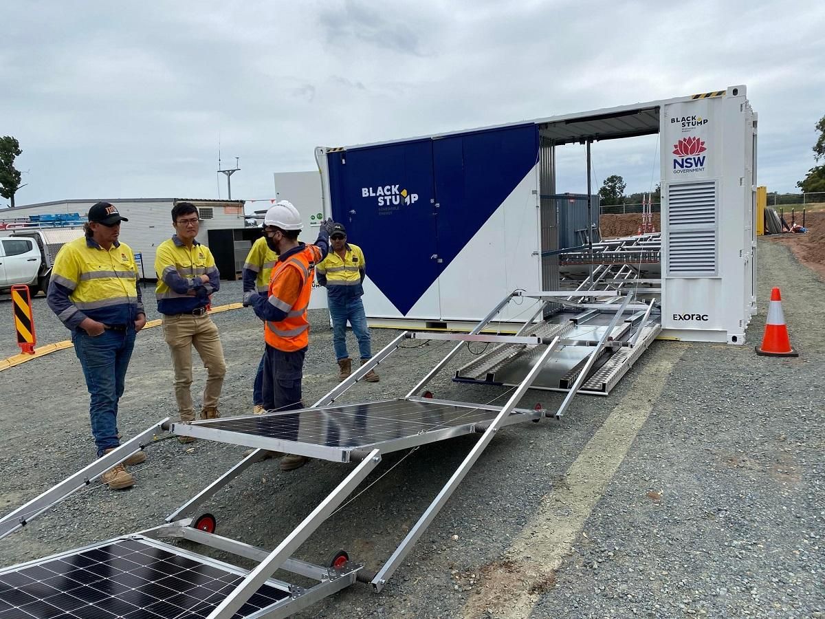 Workers examine solar panels on a ramp beside a utility container. Cloudy outdoor setting.