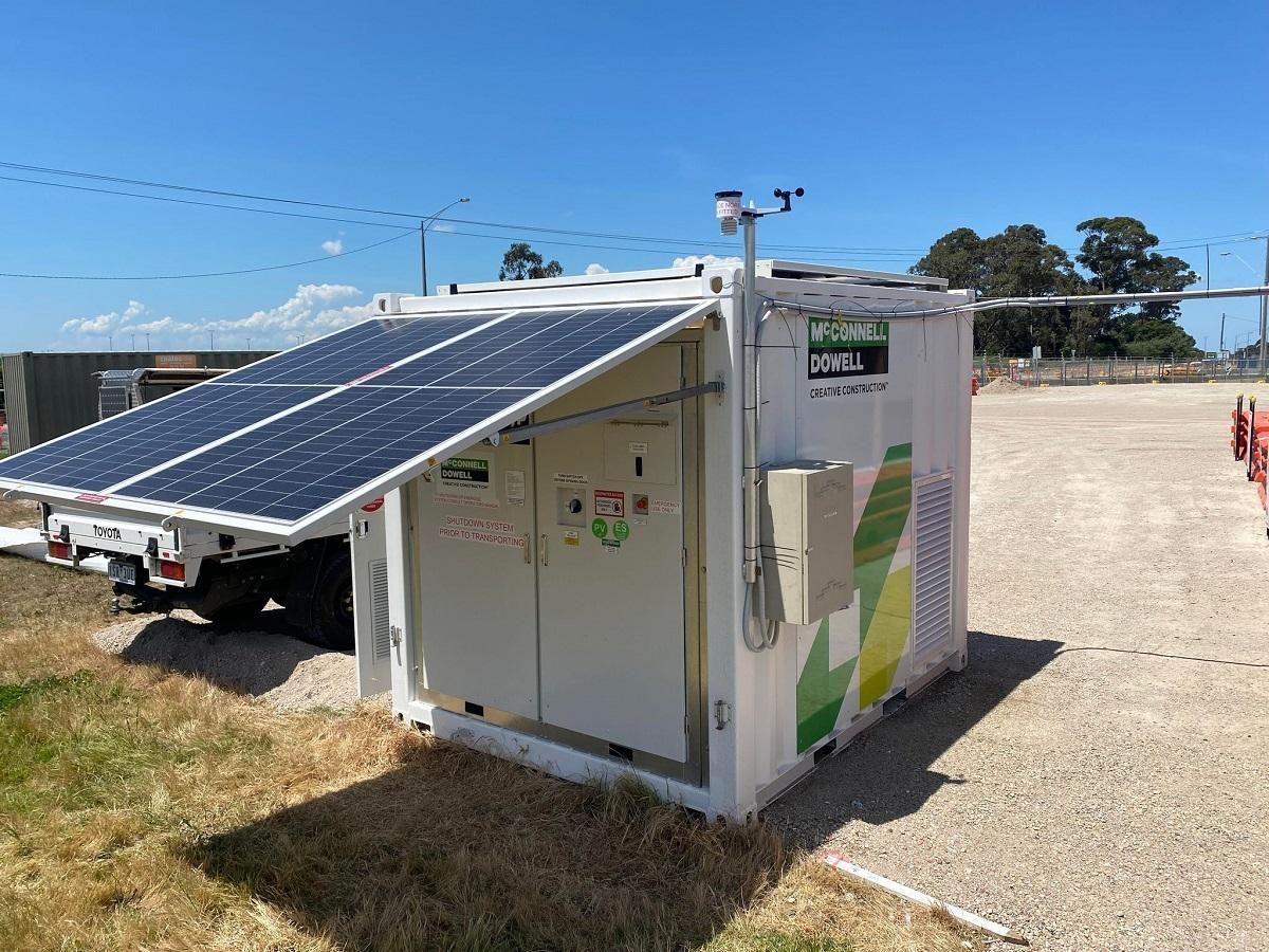 Solar panel array on a white power unit container with green branding, outdoors on gravel.