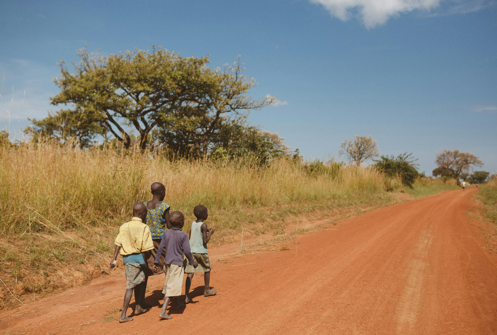 Children walking on a red dirt road, tall grass, trees, and blue sky.