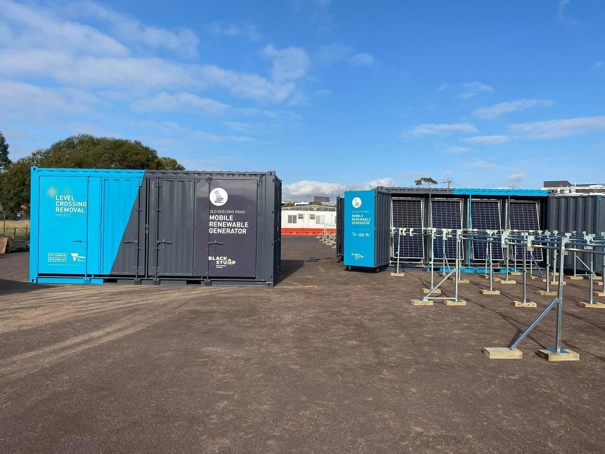 Blue and gray shipping containers with solar panels, set on a paved area, under a blue sky.