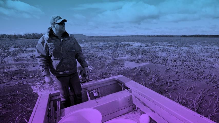 Man in a boat surveys a flooded marsh under a cloudy sky.