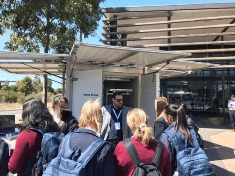 A group of students listen to a man in front of a solar-powered structure. Building in the background. Sunny day.