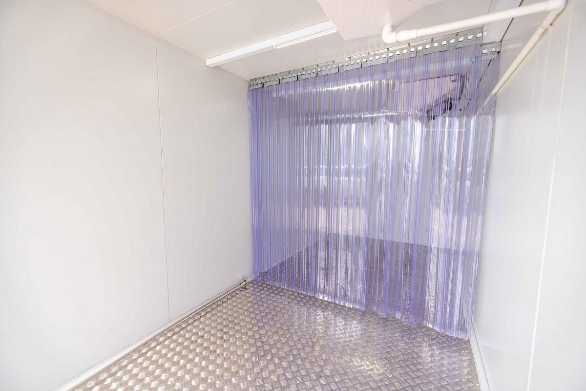 Small, white-walled room with a diamond-plated floor, plastic strip curtains, and overhead lighting.