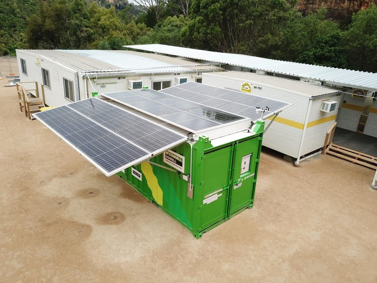 Green shipping container with solar panels, other container buildings in the background, outdoors on a sunny day.