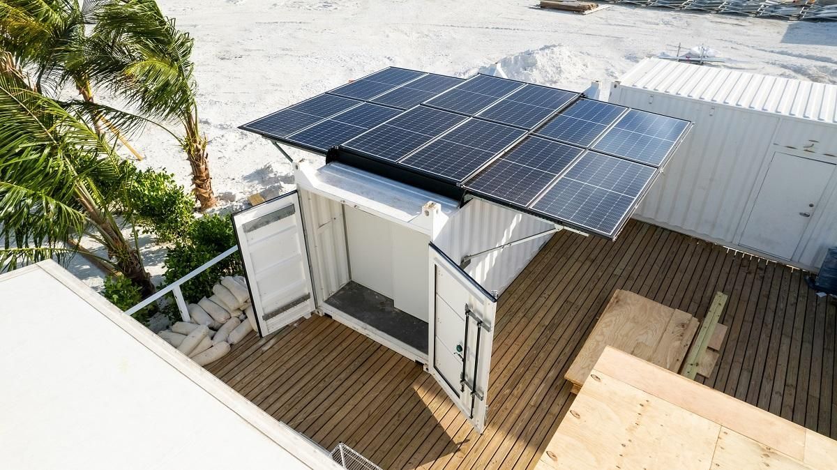 Solar panels on a white building, open doors, wooden deck, palm tree, and beach background.
