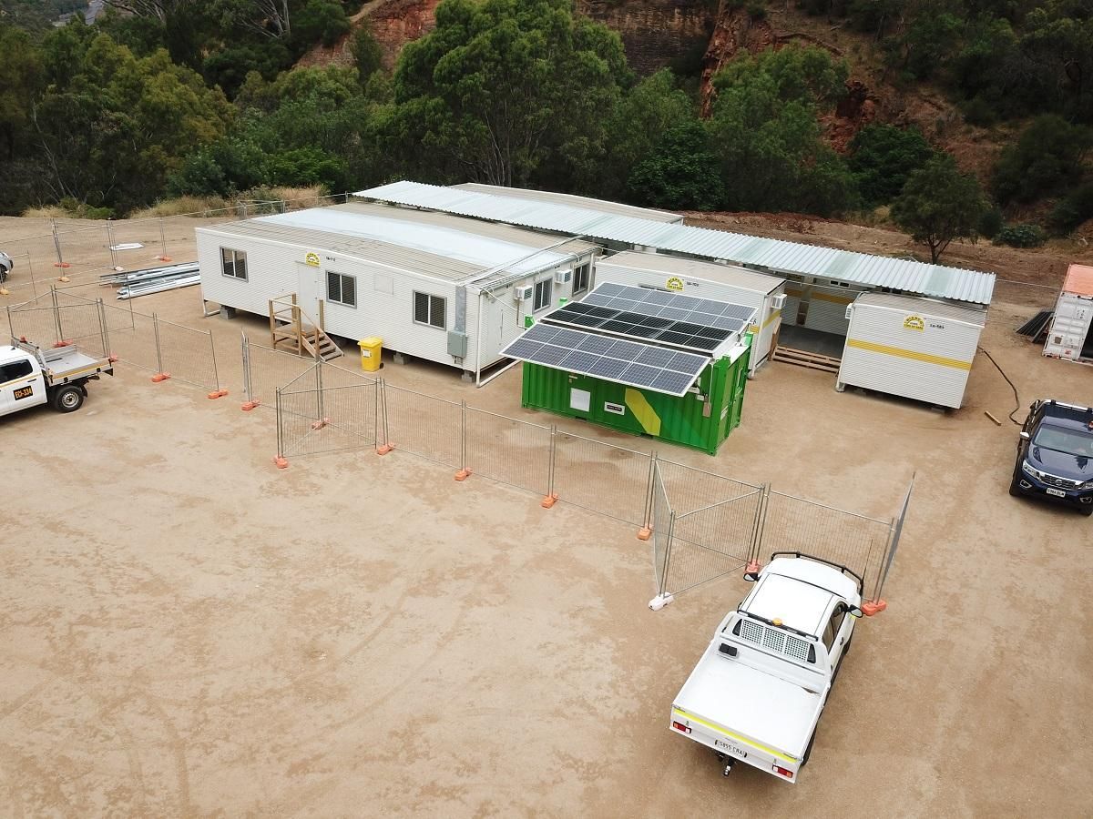 Solar-powered modular buildings on a dirt lot, with a white truck and a black vehicle parked nearby.