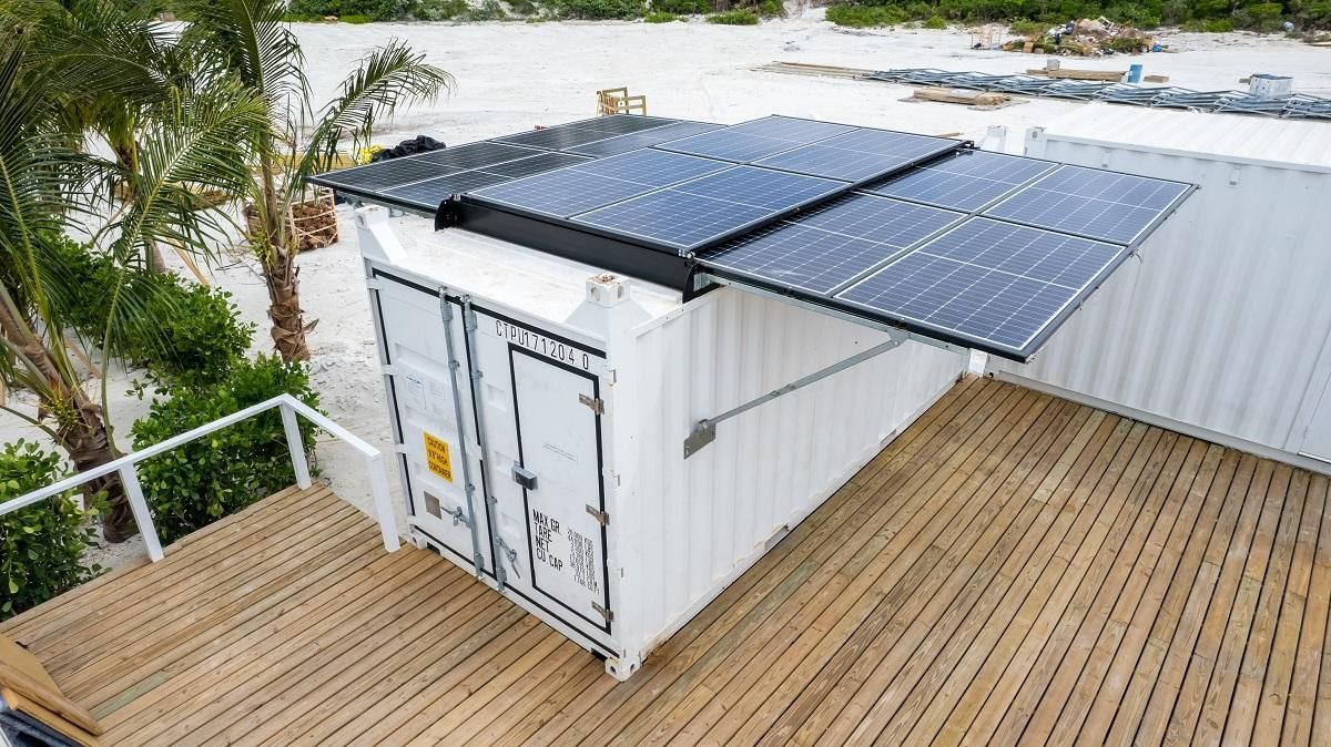 White shipping container with solar panels on a wooden deck, near palm trees and the beach.