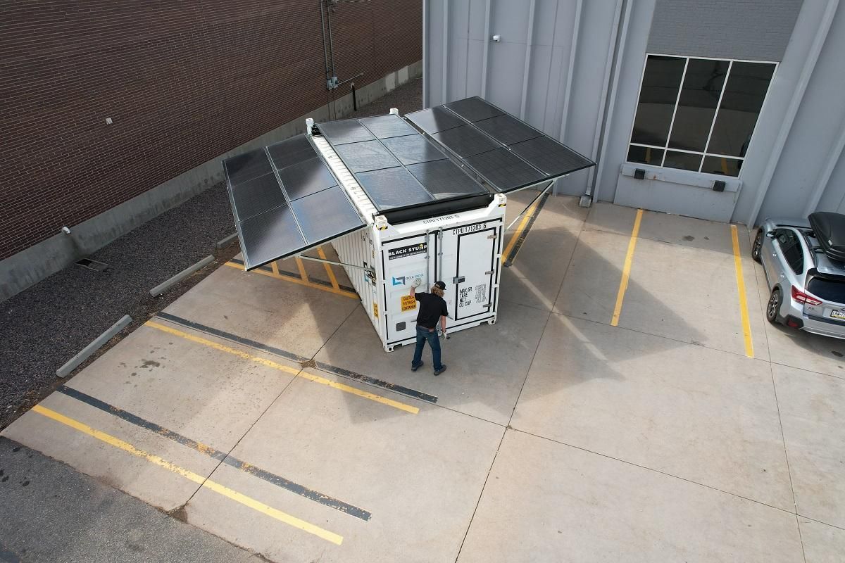 Solar panels on a shipping container in a parking lot; a person stands nearby.