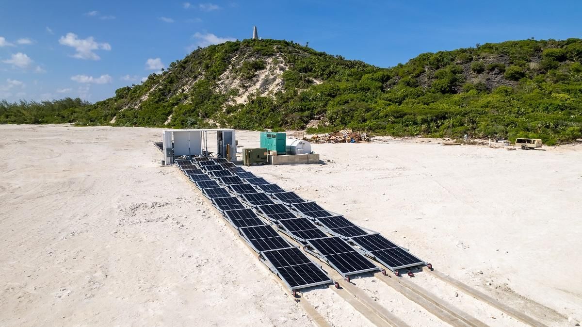 Solar panels on a sandy beach, with equipment, and a green hill in the background.