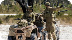 Two soldiers in camouflage gear near a beige ATV; one gestures, the other drives in a natural outdoor setting.