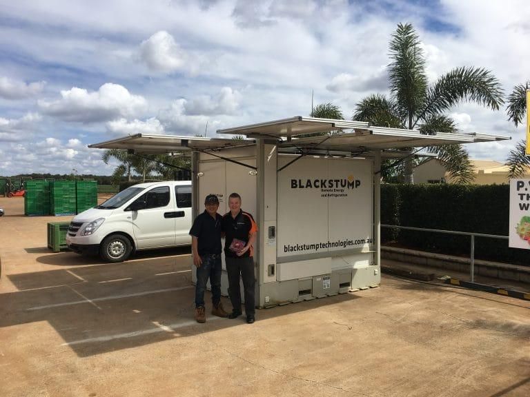 Two men stand near a white container with solar panels. A white van is parked nearby, with a blue sky.