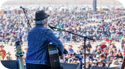 A musician playing guitar onstage at a large outdoor festival; crowd in the background.
