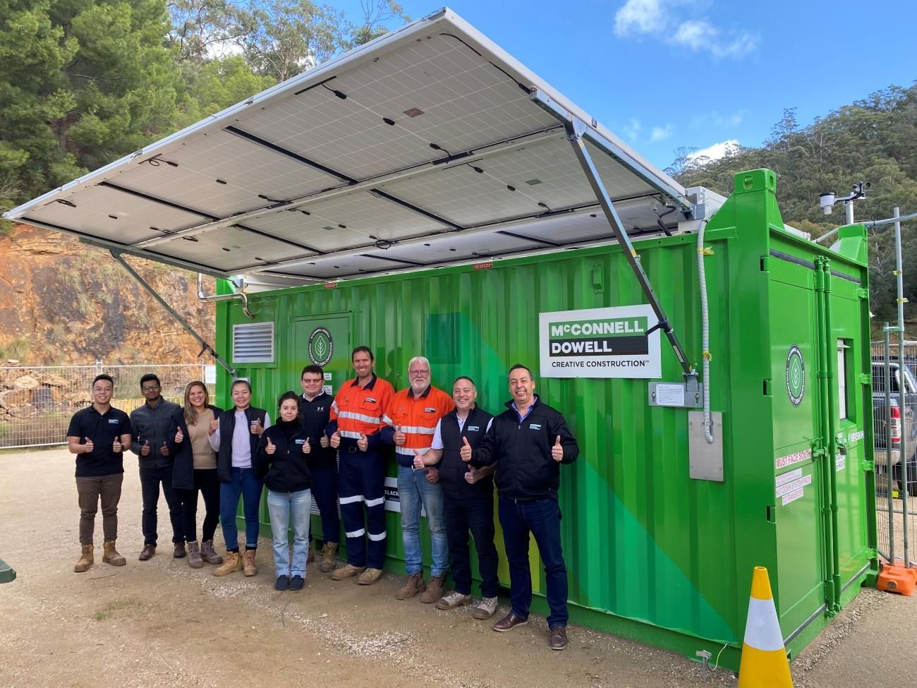 Group of people standing in front of a green container with solar panels. Everyone giving thumbs up.