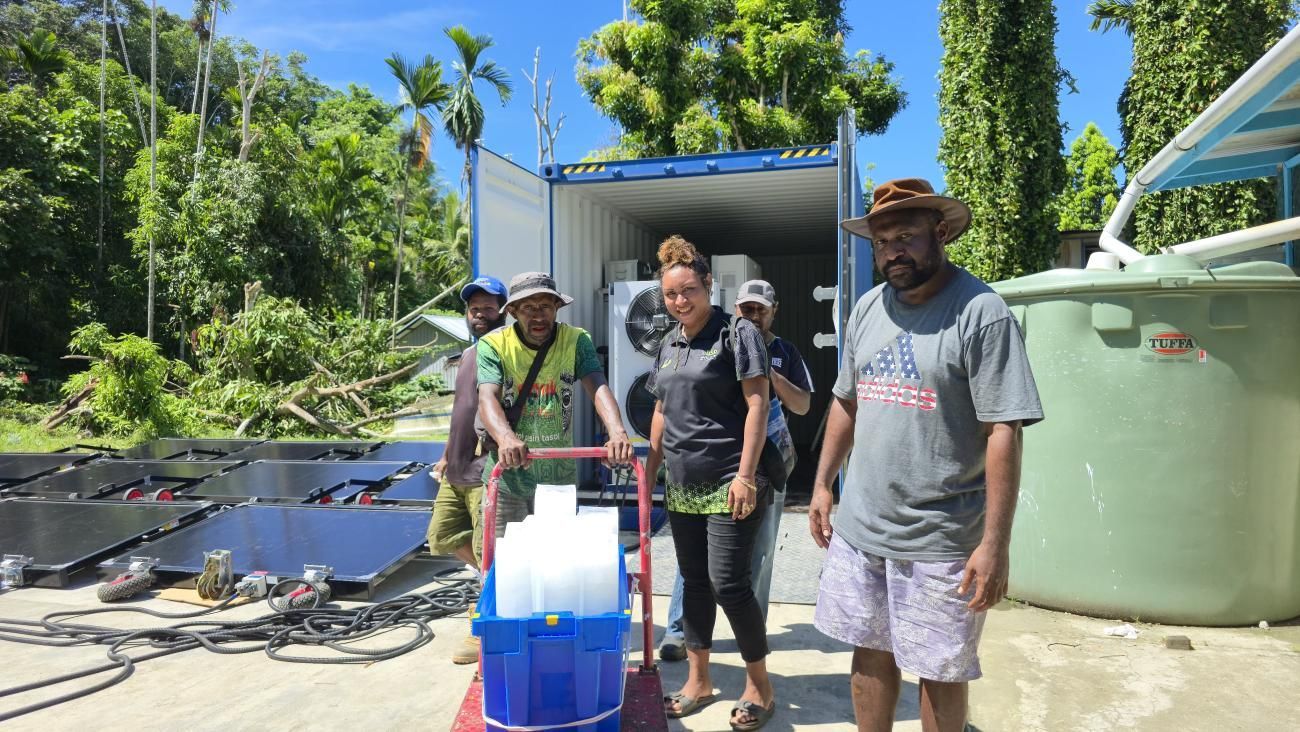 People unload supplies from a shipping container outdoors, next to solar panels and a water tank, under a sunny sky.