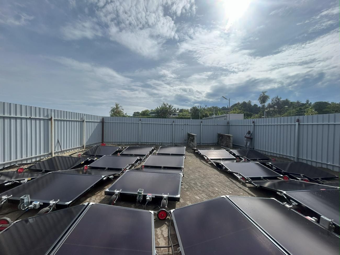 Solar panels laid out on the ground in an outdoor space, surrounded by a metal fence under a cloudy sky.