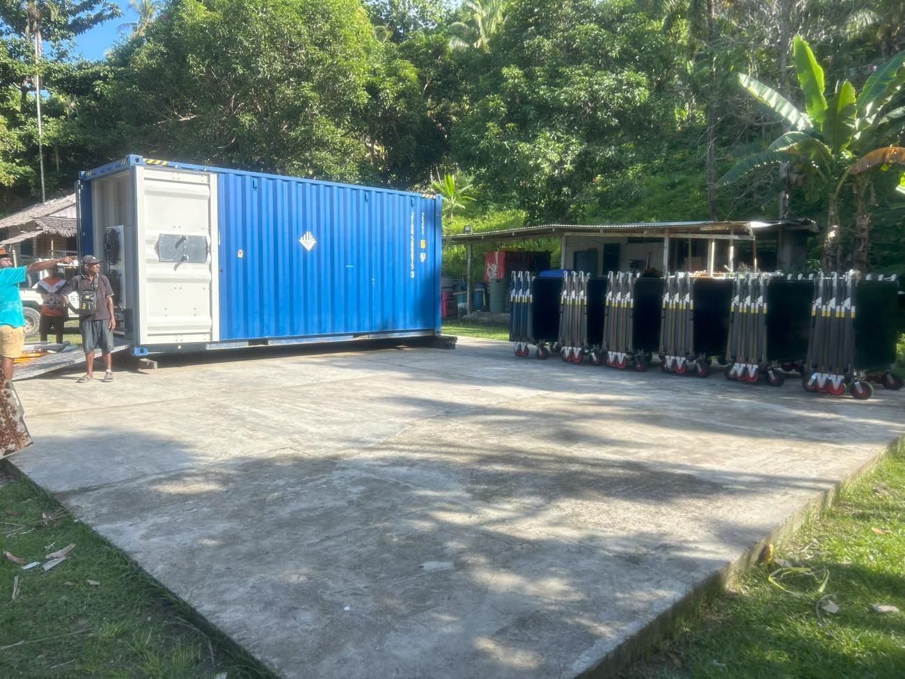 Blue container and folded equipment on a concrete pad with people nearby, trees, and a building.