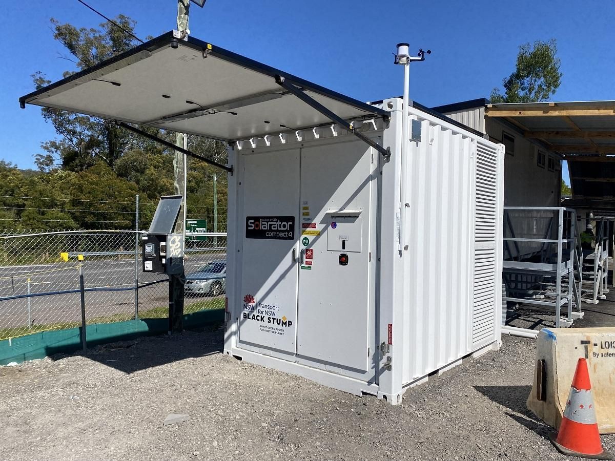 White container with solar panel roof, sensors, and a control panel, outdoors on gravel.
