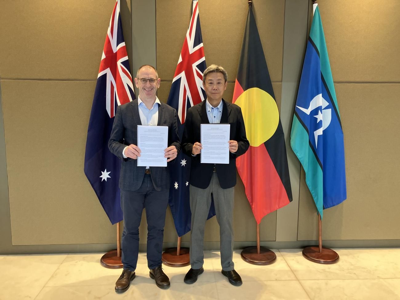 Two men holding documents, standing in front of Australian and Indigenous flags, signing an agreement.