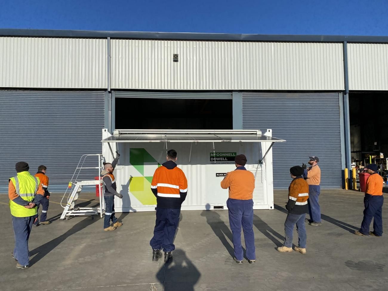 Group of people in work attire stand near a white container in front of a building with a large open doorway.