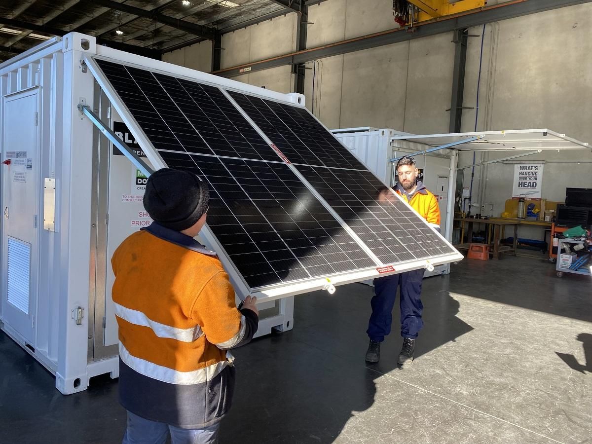 Two workers installing solar panels on a white container in a warehouse.