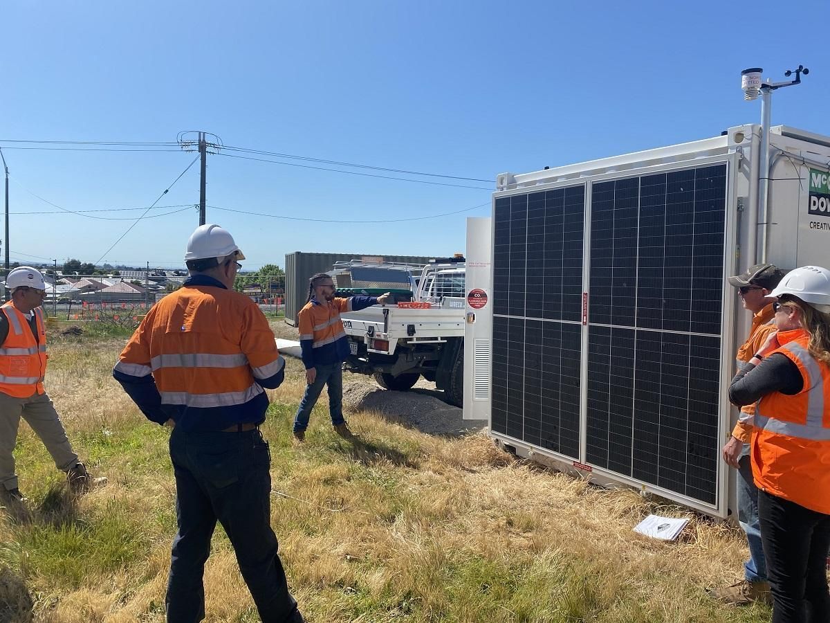 People in safety vests examine a solar panel array attached to a white container next to a work truck, outdoors on a sunny day.