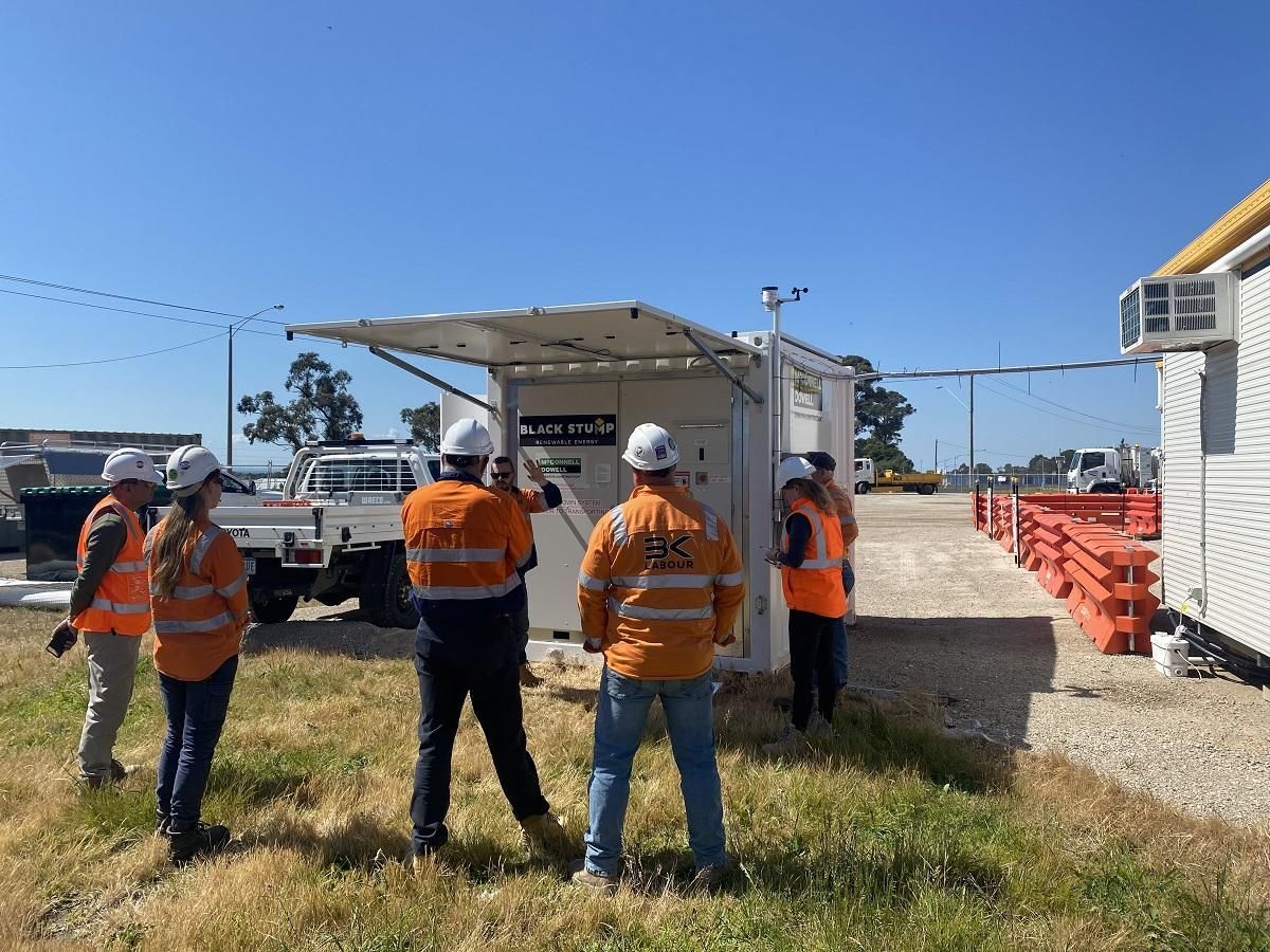 Construction workers in safety vests and hard hats examining a white control box outdoors.