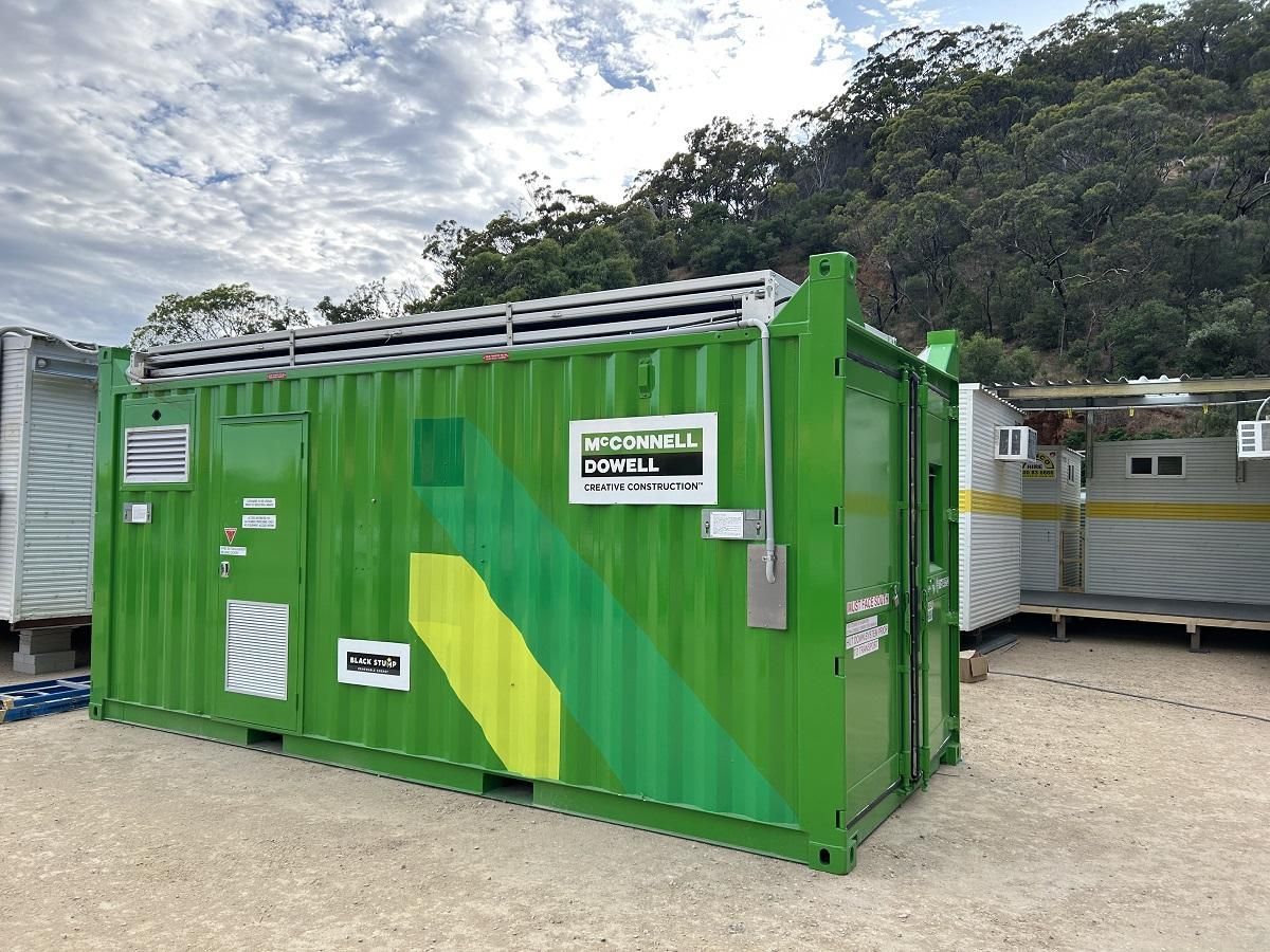 Green container with power equipment, outdoor setting, partial view of other containers and hillside.