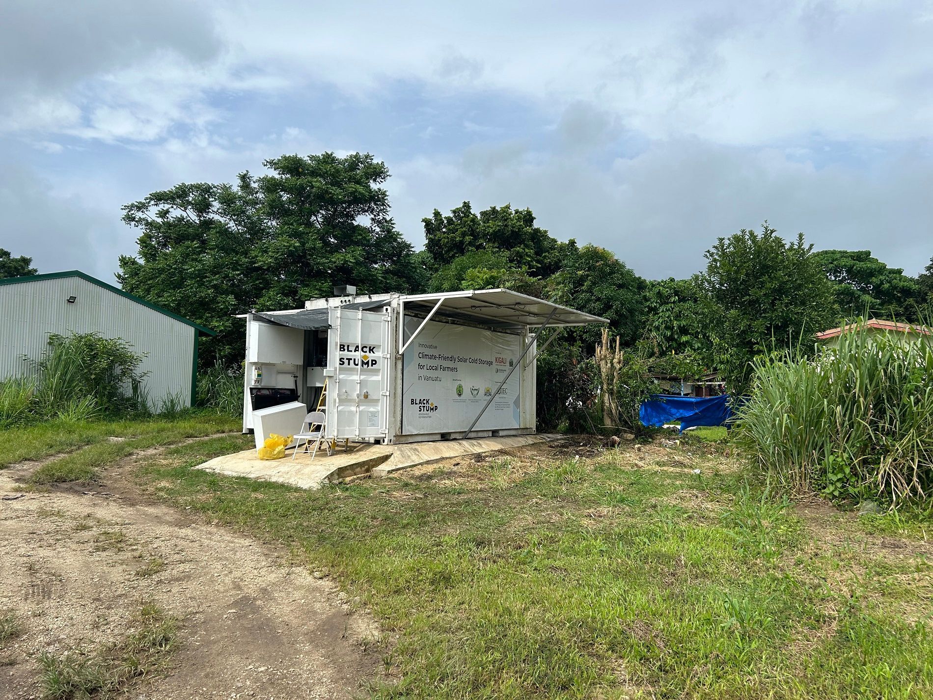 White shed-like structure with an open-air front, handwritten notes, grass, and trees under a cloudy sky.